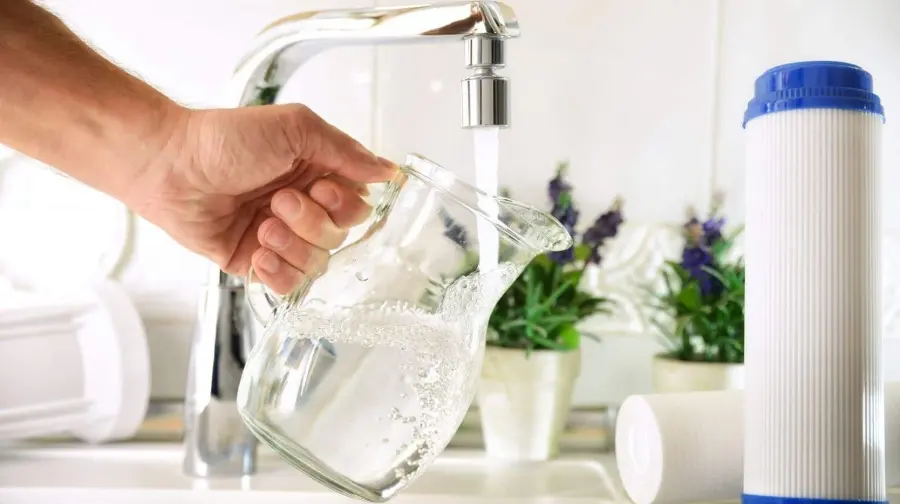 Hand filling a glass pitcher with fresh, sparkling pure filtered drinking water from the kitchen faucet, with a white and blue water filter cartridge shown in the foreground – United Water Softeners.