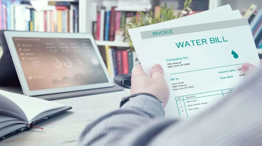 Person holding and reviewing a water bill invoice at a desk with a tablet and notebook, illustrating the financial impact of hard water on household utility costs.