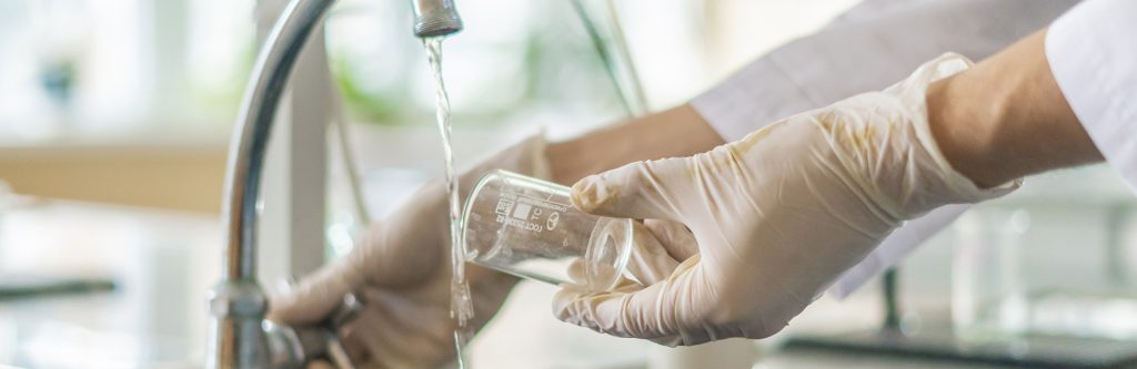 Gloved water quality technician carefully collecting a precise sample from a flowing kitchen faucet into a laboratory glass test tube – expert water analysis by United Water Softeners, Houston’s best water company
