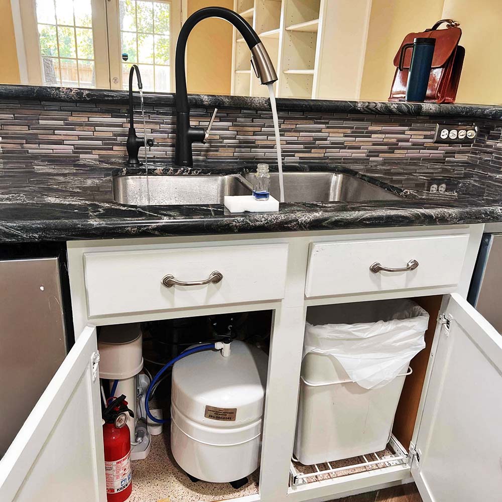 Professional PUREGUARD™ PGRO-4 reverse osmosis drinking water system installation by United Water Softeners in Spring, Texas. Custom matte black faucet on a stainless steel kitchen sink dispenses the purest, crystal-clear water into a sample bottle, with the white 4.2-gallon RO tank neatly installed under the sink.
