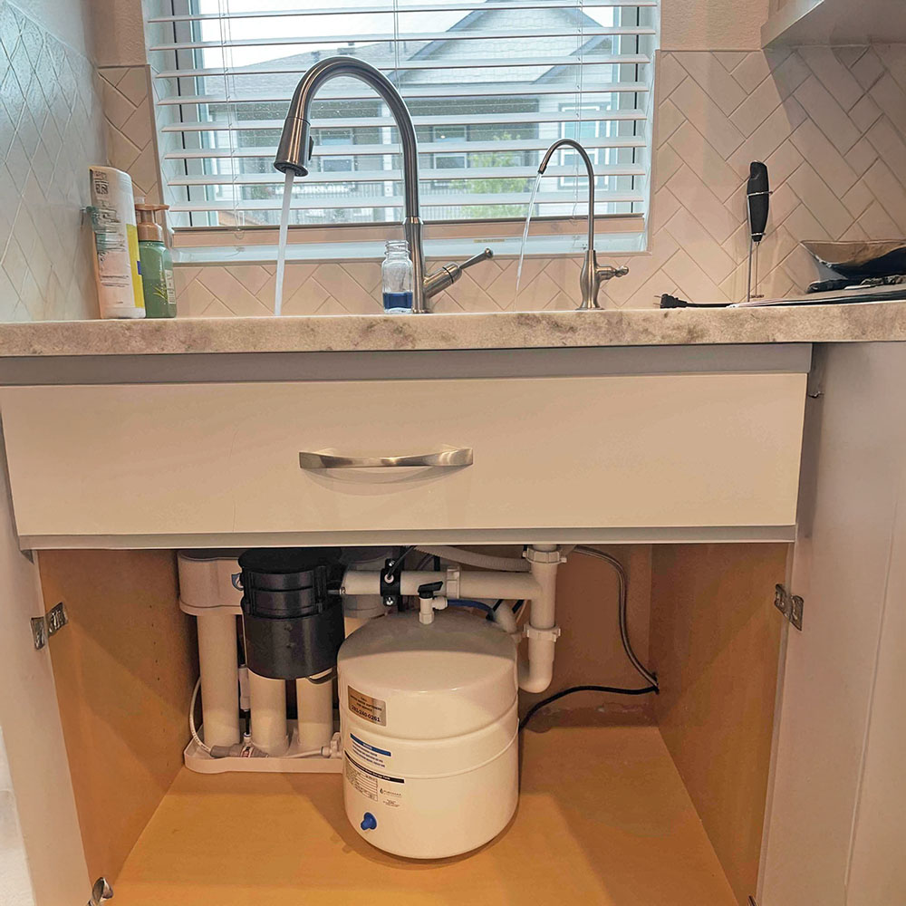 Professional PUREGUARD™ reverse osmosis drinking water system installation by United Water Softeners in Cypress, Texas. Custom chrome faucet on the kitchen sink dispenses the purest, crystal-clear water into a sample bottle, with the white RO tank and multi-stage filters neatly installed under the sink.